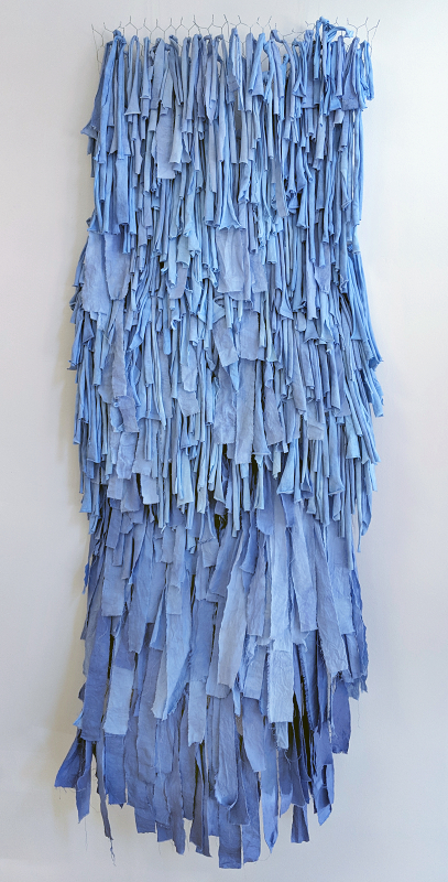A sculptural piece made of strips of fabric died in different shades of blue, tied to a background made of chicken wire. The piece is titled \"Epeme(real)\"