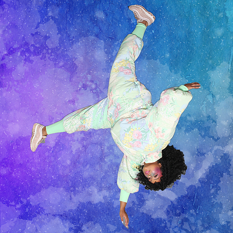 Photo of Arielle Gray wearing a space suit made out of an old floral comforter. The artist floats upside down over a blue and purple speckled background meant to resemble space.