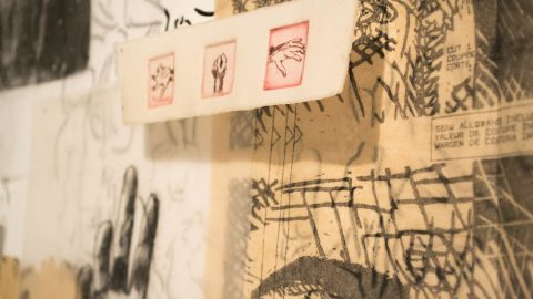 Pieces of paper with black and red pen sketches of bodies and body parts are layered on a wall using small pins