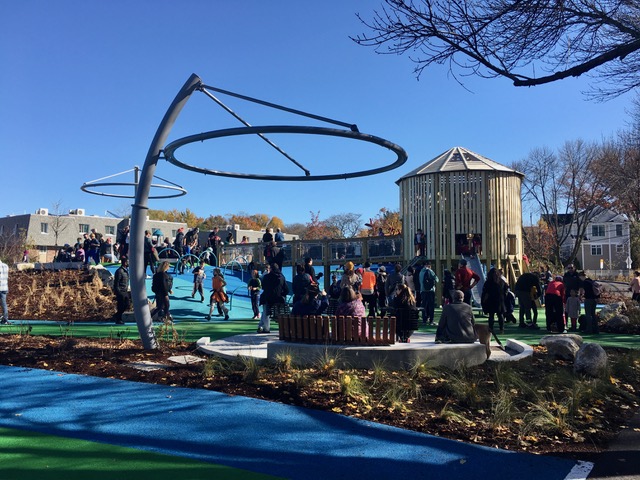 Image of playground with blue and green rubber ground and many people standing wearing jackets to celebrate the opening