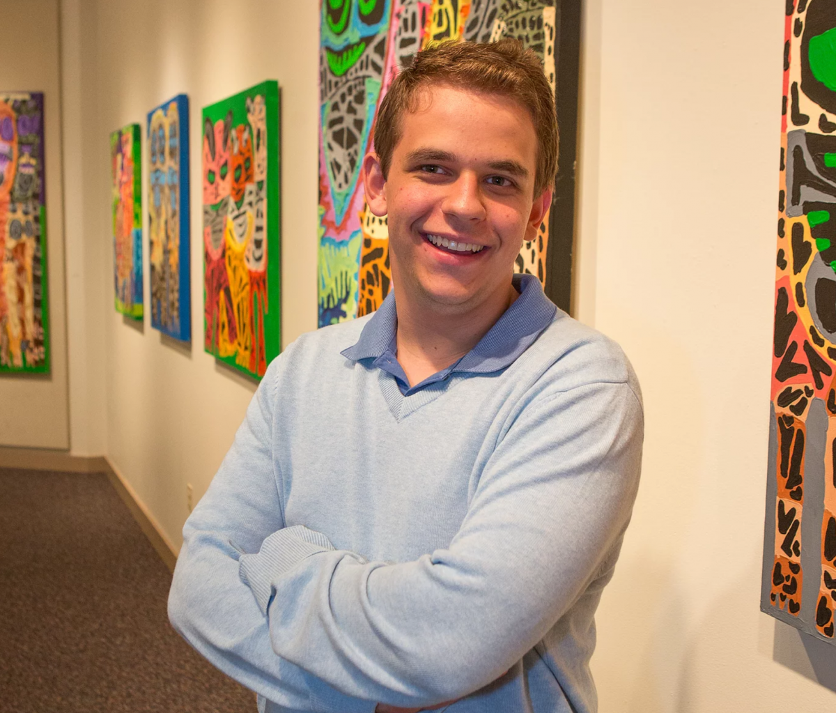 Image of Dominic smiling with his arms crossed, standing in front of his work hanging in a gallery