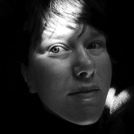 A black and white portrait of Megan Bent. She has pale skin and dark bangs sweeping over her forehead. Most of her face is in a light shadow and the sun illuminates one eye