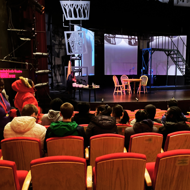 a photo of the back of students heads in red theater chairs facing an empty stage set with a table and chairs