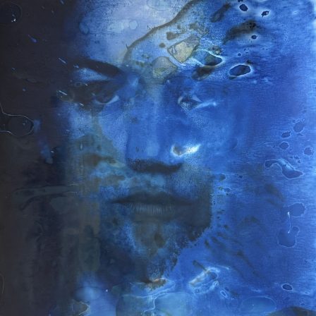 The self-portrait is rendered in a deep indigo blue. On the right is a large watermark in light blue. It almost appears to wash away the right side of her face. In the watermark are green splotches. The watermark and green splotches are from chemicals being added to the cyanotype print.
