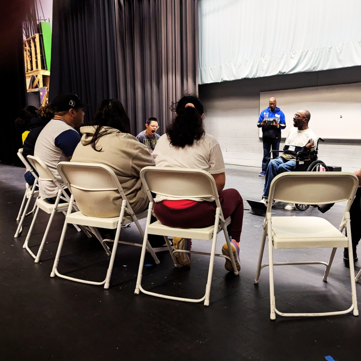 a photo of students sitting in white folding chairs in a circle featuring a speaker, Keith Jones, a black man using a motorized wheel chair
