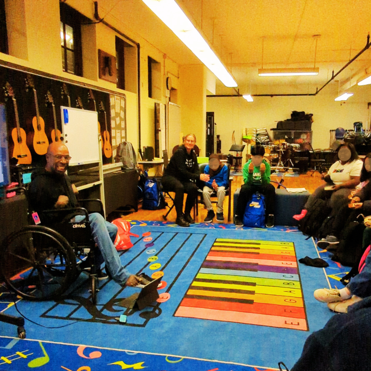 a group of students sit in a circle facing speaker, Keith Jones, a black man using a motorized wheelchair, smiling in front of several hanging guitars