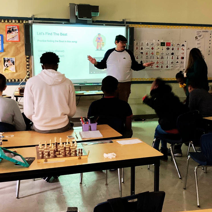 a group of students sit on tables and chairs facing speaker, Mehdi Raoufi in front of a smart board featuring music content