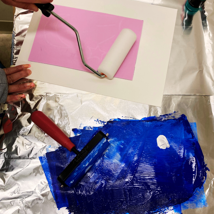 a photo featuring hands holding two paint rollers, one rolling blue paint onto a print