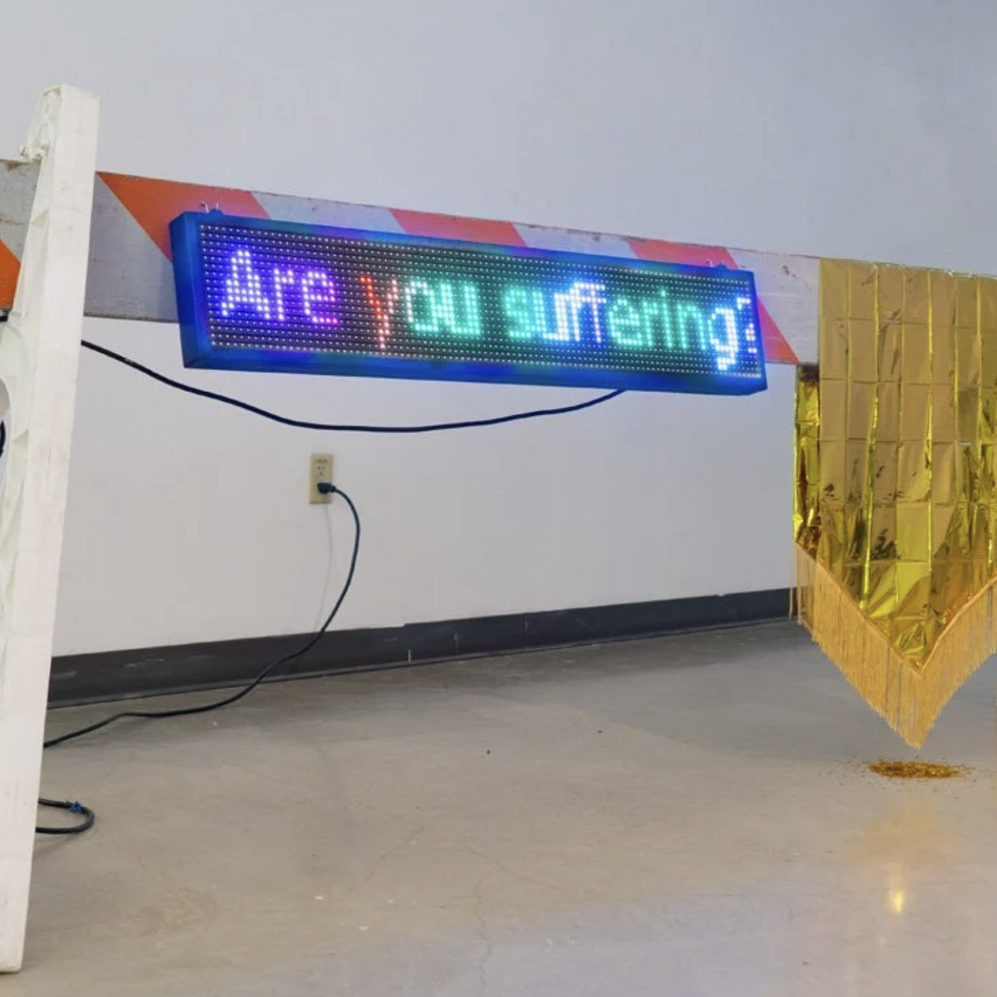 A large neon orange and white road blockade features an LED screen with the words "Are you suffering?" On the right side of the blockade is a shimmery gold emergency blanket and a pile of gold confetti underneath it.