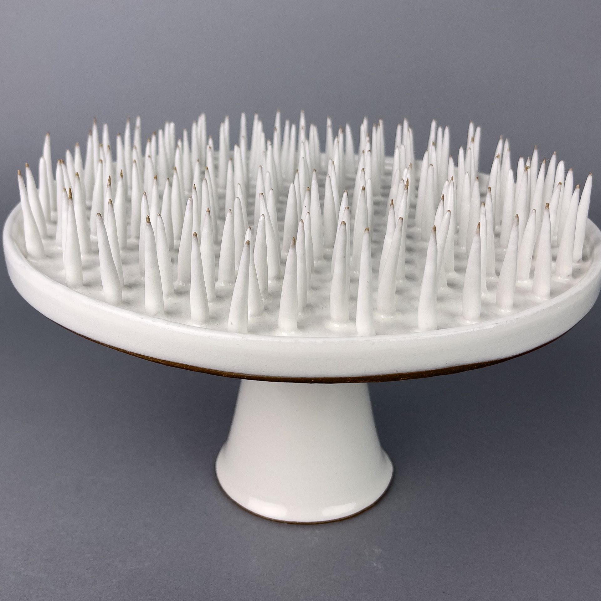 Hostile Cake Stand by Hayle Lovstedt A white ceramic cake stand. The top surface of the cake stand is covered in long, white, pointy spikes.