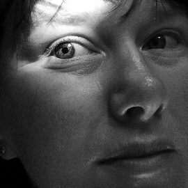 A black and white portrait of Megan Bent. She has pale skin and dark bangs sweeping over her forehead. Most of her face is in a light shadow and the sun illuminates one eye