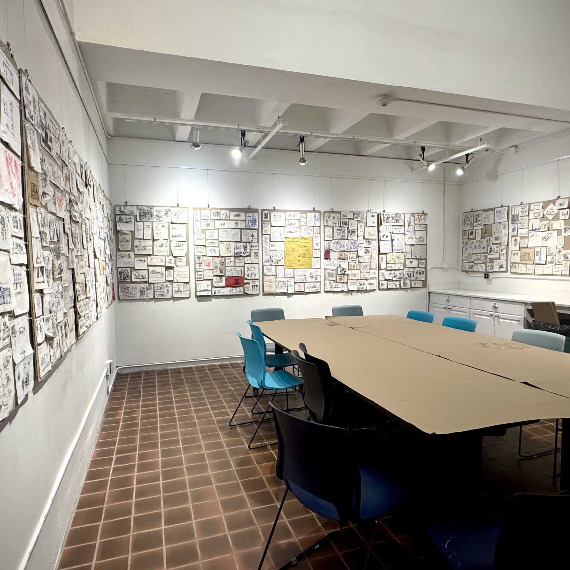 A salon-style installation of Tim’s napkin sketches. These sketches were made by Tim over the last 5 decades. The napkins are pinned to large cardboard panels that cover the surface of the three walls in the studio. The napkins are various sizes and colors. Some napkins are square, some rectangular, and some have drawings on both sides, so they have been displayed unfolded. The ink colors also vary from blue, black, green, red, and orange. The subject matter is predominantly sketches of ideas present in Tim’s art, like buildings, boats, people together, and dogs. There are also written reflections, resources, and to-do lists. The display of the napkins together feels like a generous offering from the artist, him opening up his visual diary for us to peruse.